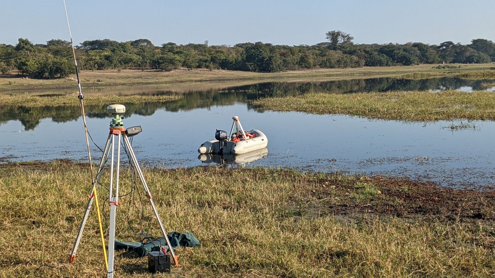 GNSS base station on a tripod on the shore, with the survey boat on the water behind it